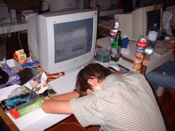 A person with their head down on a cluttered desk in front of an old CRT monitor, surrounded by snack bags, soda bottles, and crumbs.