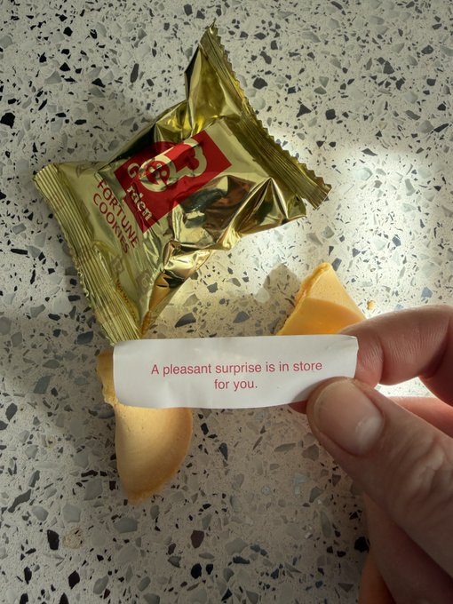 A hand holds a Lotus brand fortune cookie paper reading 'A pleasant surprise is in store for you.' above the broken cookie pieces and gold wrapper on a speckled countertop.