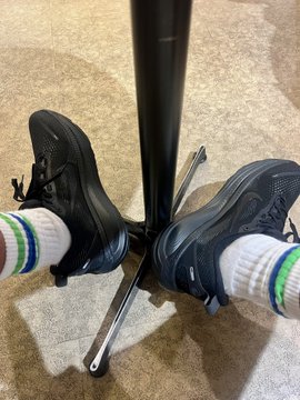 The image shows a person's feet wearing black Nike sneakers, with white socks underneath them. The shoes are positioned on a metal stand or pole, which appears to be located indoors. There is also a green sock visible near the bottom of the frame. The background seems to be a room where the person has placed their shoes and socks for easy access.