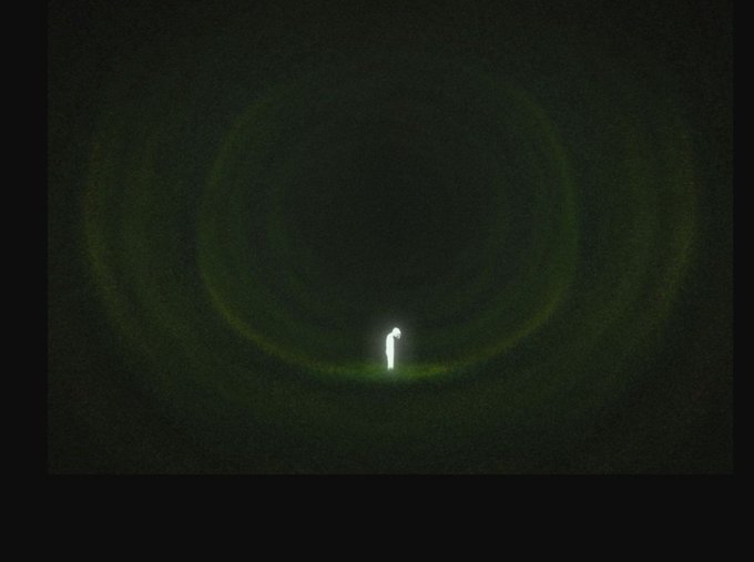 A minimalist image featuring a glowing lowercase 'i' surrounded by a greenish halo against a dark, grainy background.
