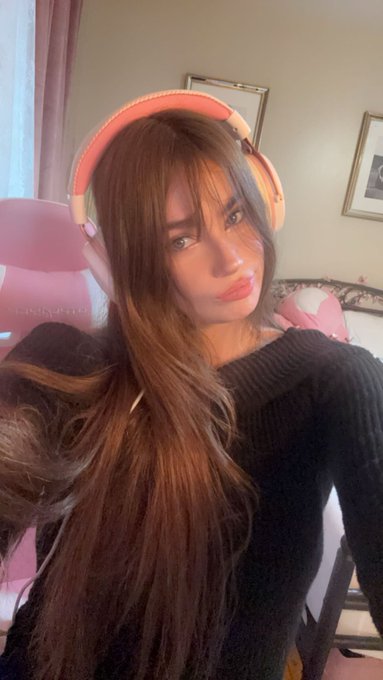A young woman with long brown hair wearing pink over-ear headphones poses for a selfie in what appears to be a cozy bedroom, giving a casual "yo" greeting vibe with her relaxed, warm-toned setting.