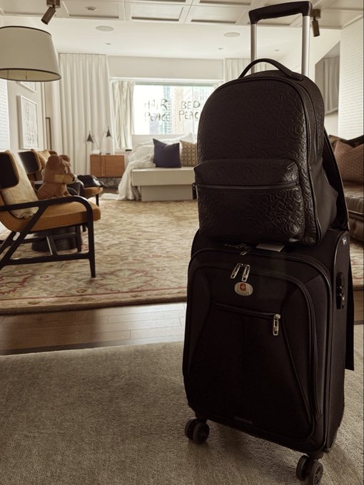 A black leather SwissGear rolling suitcase with a matching backpack attached sits on a carpet in a hotel room living area with chairs and a teddy bear in the background.