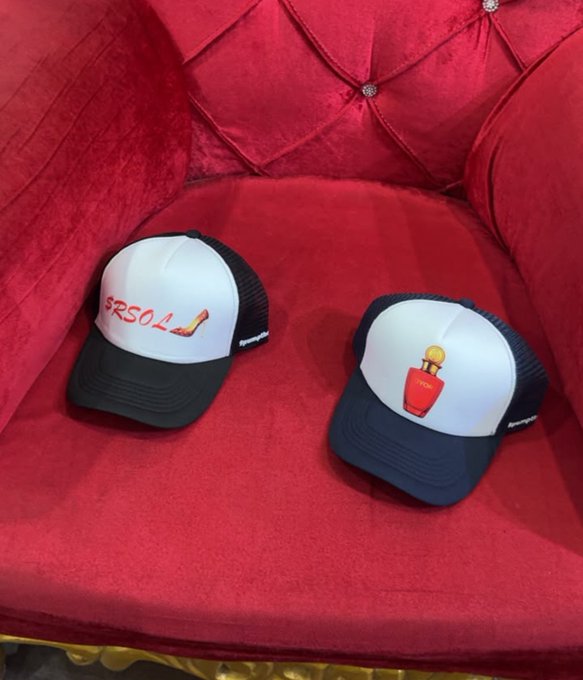 Two black and white trucker hats sit on a red tufted chair; one features a 'PERSOL' logo with a shoe graphic, while the other displays a perfume bottle graphic on the front panel.