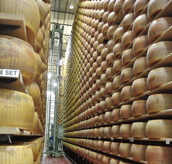 Rows of aging Parmesan cheese wheels, including one marked "4 SET," tower on tall racks in a storage facility, visually emphasizing the traditional, long-term aging process that mirrors the "stablecoin" theme of reliability and value preservation over time.
