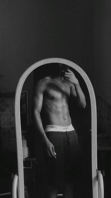 A black and white mirror selfie showing a shirtless man wearing dark pants, holding a phone to take the photo with his face obscured by the camera.