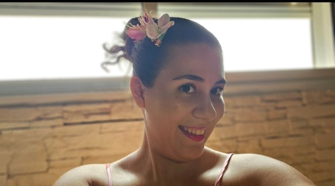 A portrait of a smiling woman with dark hair styled in a bun adorned with pink and white flowers, wearing a pink top against a stone wall background.