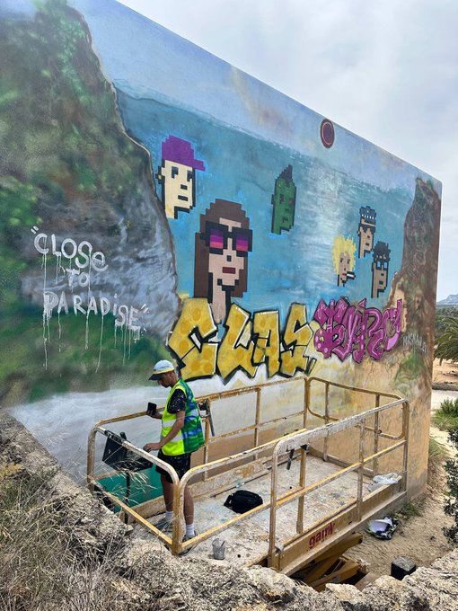 A worker in a yellow safety vest operates a scissor lift in front of a large outdoor mural featuring pixel-art characters and graffiti text including 'Close to Paradise' and 'LEGS'.