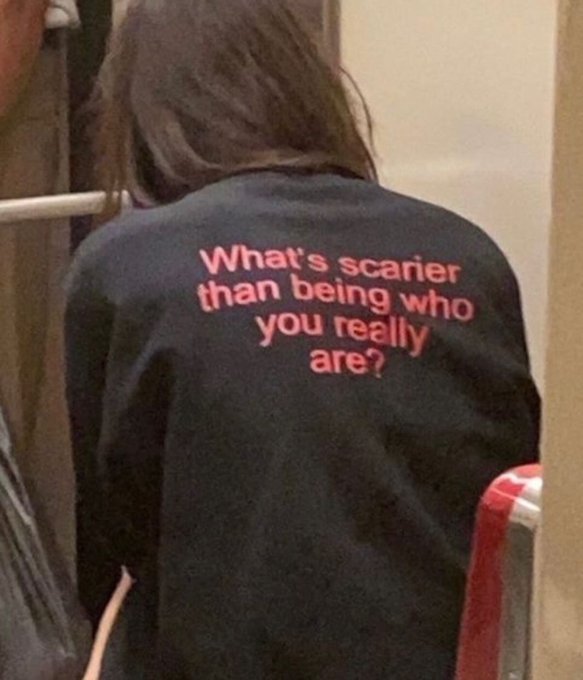 A person with long brown hair, photographed from behind, wears a black shirt featuring pink text that reads "What's scarier than being who you really are?"