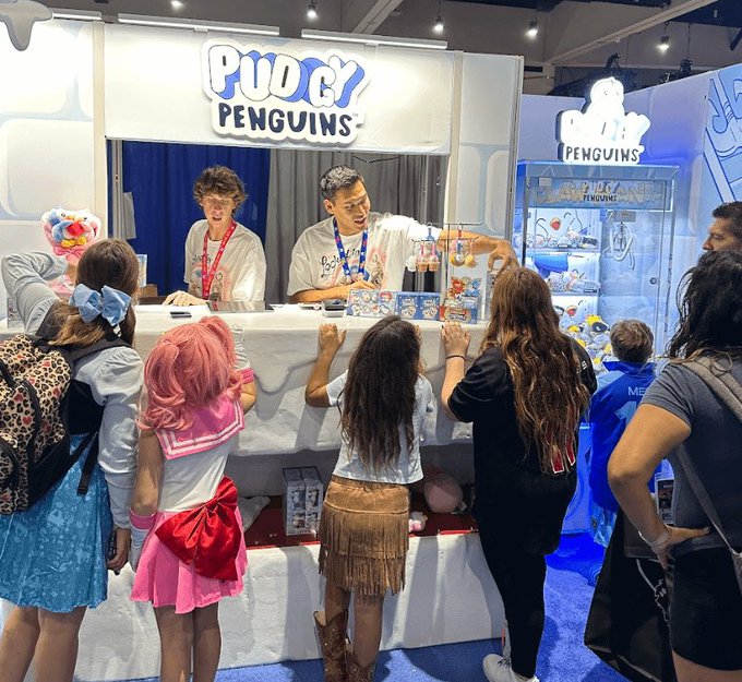 Fans gather at a 'Pudgy Penguins' booth, likely at a convention, where two staff members interact with attendees including one person dressed in a pink cosplay outfit.