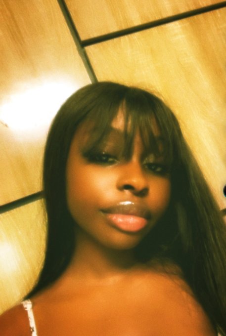 A close-up selfie of a young woman with long black hair, bangs, and glossy lips, wearing a white spaghetti-strap top, taken against wooden paneling with warm lighting.