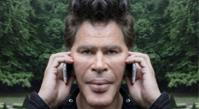 A man with spiky hair holds two mobile phones up to his ears, set against a forest background.