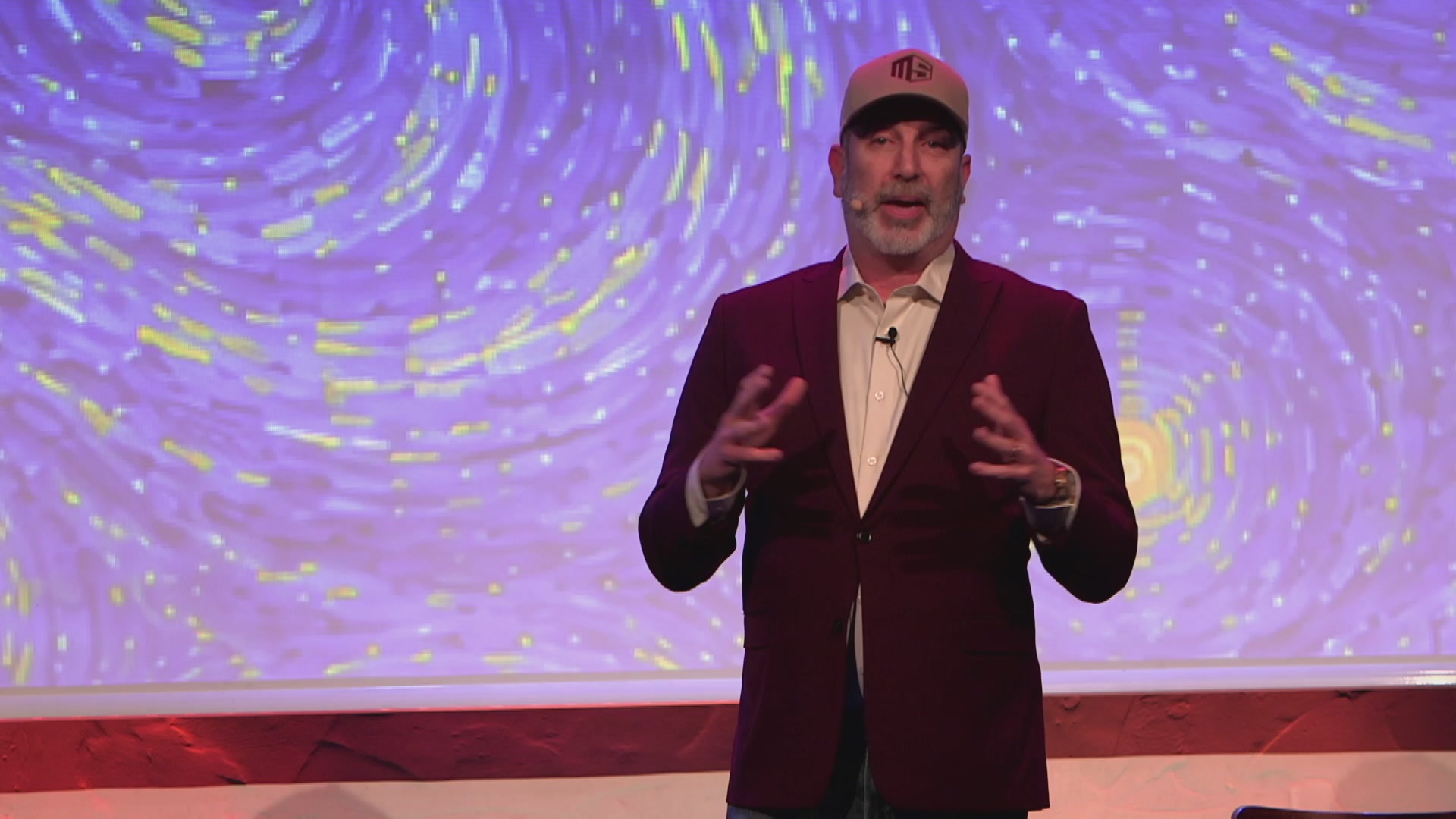 The image features a man wearing a red suit, standing behind a podium with his hands outstretched as if he's giving a speech or presentation. The background of the image shows a large screen displaying a starry night sky, which appears to be part of an art installation or multimedia display related to the event taking place at the Litecoin Summit.

The caption on the image reads "The Litecoin Summit is live beaches! Come join us for Day 1." This suggests that the man in the suit might be addressing a crowd about Litecoin, possibly during a panel discussion or presentation.