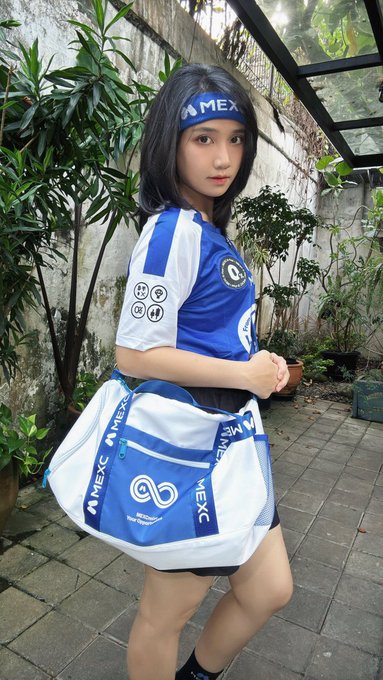 A young woman poses in an outdoor courtyard wearing a blue and white jersey and matching headband branded with "MEXC" logos, carrying a coordinating duffel bag that also displays the MEXC name alongside an infinity-style emblem, perfectly matching the post's enthusiasm for the limited World Cup edition merchandise.