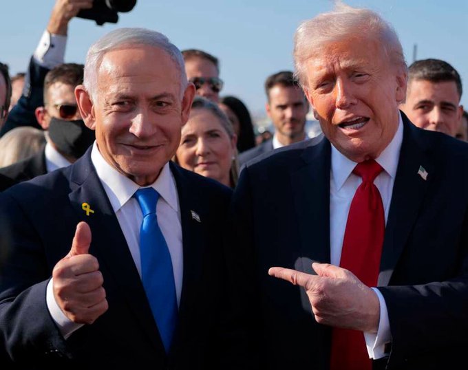 The image shows two men, possibly former presidents or political figures, standing next to each other and giving each other a thumbs up sign. They are both dressed in suits and ties, with one of them wearing sunglasses. The background includes several people who appear to be watching the interaction between the two men.