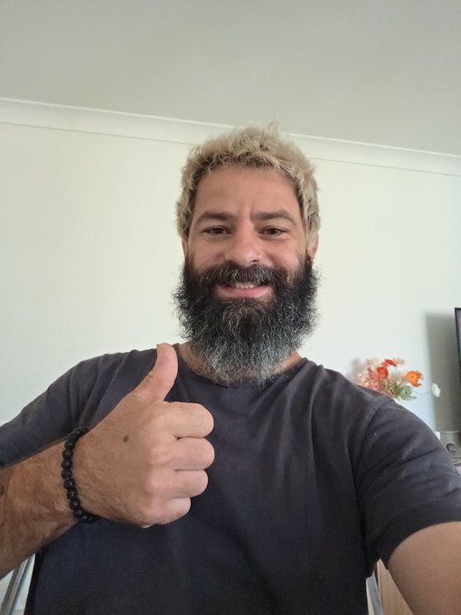 A man with a grey-streaked beard and blonde hair smiles and gives a thumbs-up while wearing a dark t-shirt.