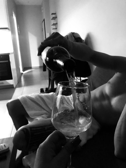 A shirtless person relaxes on a couch while pouring white wine from a bottle into a glass, capturing a casual "good night" moment that pairs with the social post tagging @River4fun and @RiverdotInc with the ticker $RIVER.