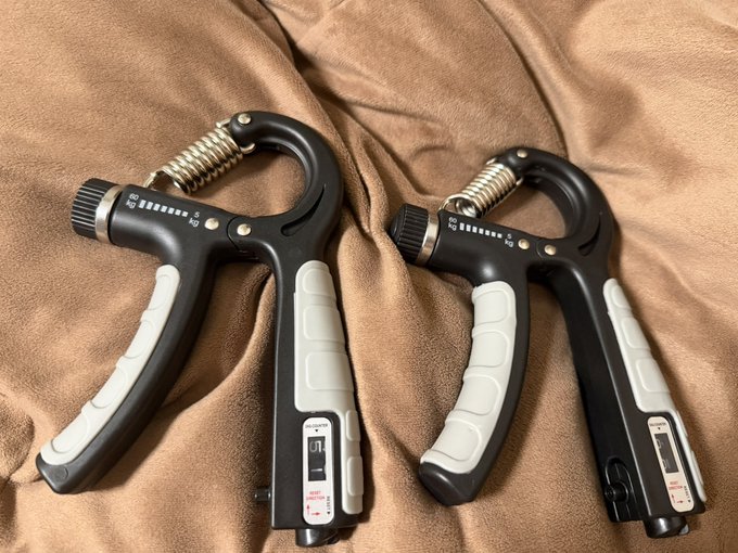 A pair of black and white hand grip strengthener exercise devices with adjustable dials and metal springs, resting on a tan fabric surface.