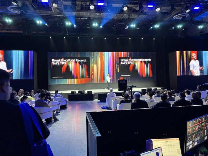 A conference or tech event with a speaker on stage presenting 'Break the World Record: Sui's TPS' on multiple large screens to an audience seated in a large auditorium.