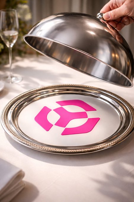 A hand lifting a silver cloche off a platter to reveal a pink geometric logo on a white surface, set on a dining table.