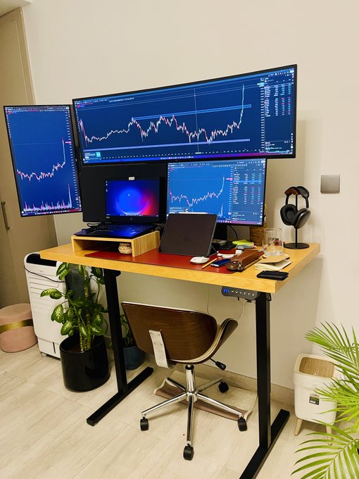 A home trading setup featuring four monitors displaying financial charts, a laptop, a laptop stand, and a modern chair in a room with plants and a wooden desk.