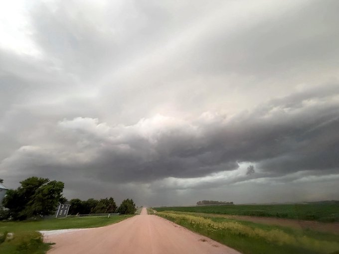 The image shows a road running through a rural area with green grass, trees, and fields. The sky above has dark clouds, suggesting an overcast day or possibly an approaching storm. The photo appears to have been taken from inside a car on the side of the road, capturing the view of the landscape as it passes by.