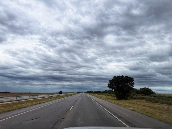 A view from inside a vehicle driving down a long, empty highway under an overcast, cloudy sky with flat plains and a single tree on the right.