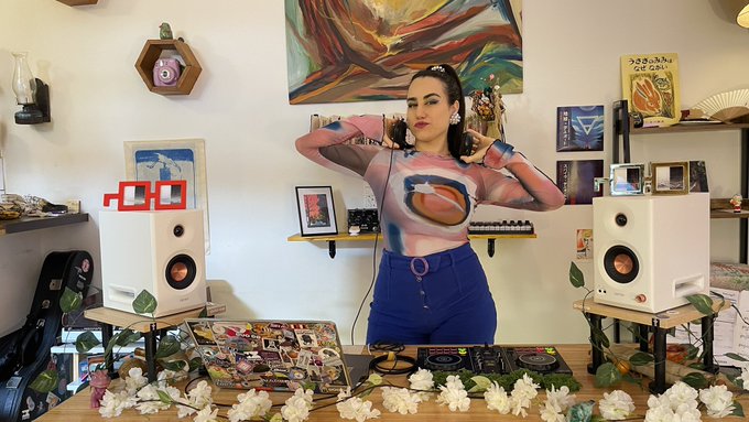 The image features a woman standing behind two speakers, wearing a unique shirt with an abstract design that includes a face painted on her chest. The background of the photo shows various items such as books, flowers, and pictures, creating a creative and artistic atmosphere. The caption for the image reads "What a fun experience it was to record my first DJ SET for @decentraland Now you can enjoy it at home too!".