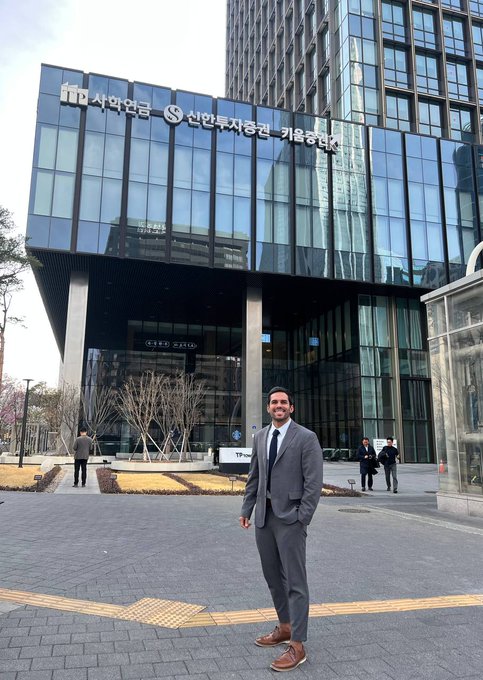A man in a gray business suit and tie posing in front of a Shinhan Investment building (서민은) with Korean signage and modern glass architecture.