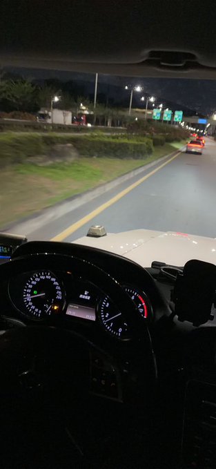 A nighttime view from inside a car driving down a highway, with the illuminated dashboard and steering wheel visible in the foreground and another car's taillights ahead. An open book rests on the dashboard, serving as the "Codex" keeping the lonely driver company on the empty road.