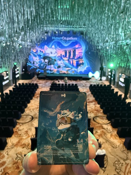 A person holds up a "Xulian" trading card at a HypurrCo gathering event, with the stage backdrop reading "HypurrCo gathers" behind rows of empty seats. The image captures the moment of pulling this sought-after grail card at the venue.