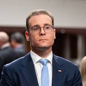 A man wearing glasses, a dark blue suit, and a light blue tie with an American flag pin, looking serious in what appears to be a courtroom setting.