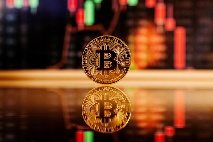 A gold Bitcoin coin stands upright on a reflective surface, with a blurred stock or crypto trading chart featuring red and green candlesticks visible in the background. The image visually reinforces the post's theme of Bitcoin projects and cryptocurrency markets, though the security vulnerability aspect is not depicted directly.
