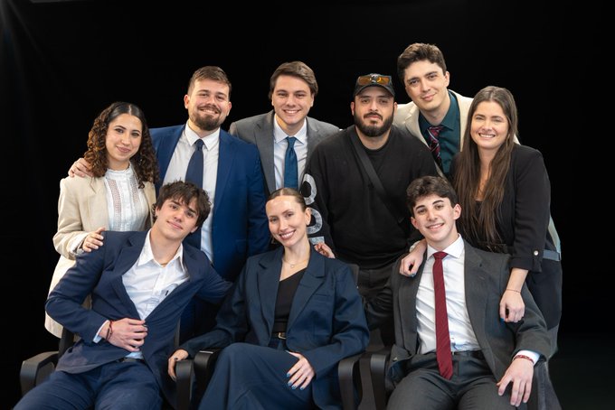 A group photo of eight people, dressed in formal and semi-formal attire, posing closely together against a dark background.