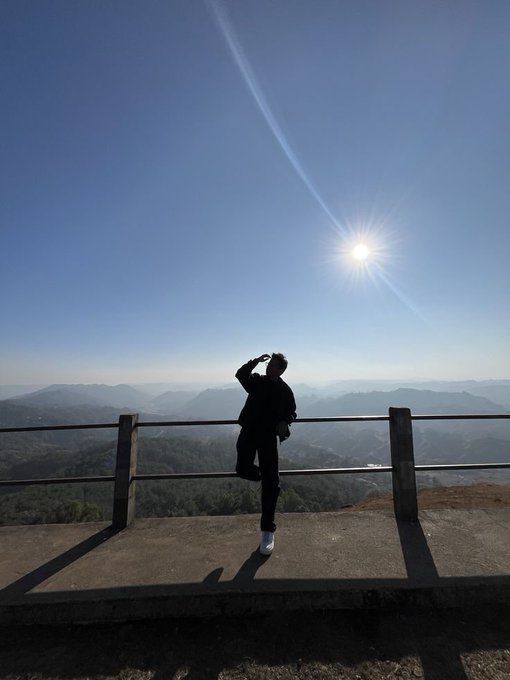 A silhouette of a person standing on a mountain overlook platform, shielding their eyes from the bright sun against a backdrop of distant mountains.