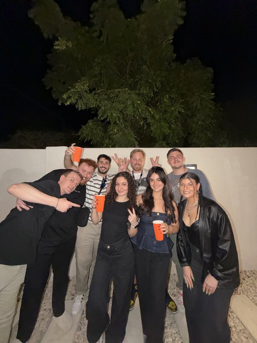 Group of eight young people posing together outdoors at night, holding orange cups and making peace signs.