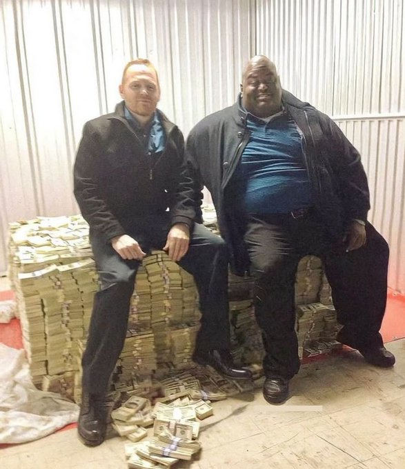 Two men in dark jackets pose sitting on massive stacks of banded US currency bills inside what appears to be a storage container or warehouse.