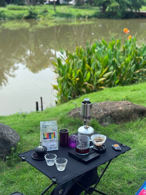 An outdoor coffee brewing setup on a black folding table by a lake, featuring a glass coffee server, metal kettle on a burner, and pour-over equipment.
