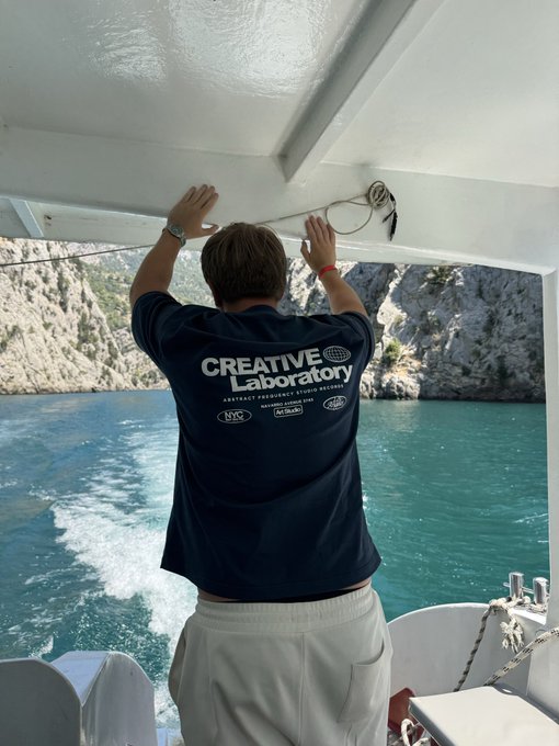 A photograph taken from behind of a person wearing a dark blue 'CREATIVE Laboratory ABSTRACT FREQUENCY STUDIO SHIRT' and white shorts, standing on a boat with their hands on the ceiling, looking out over turquoise water toward a rocky coastline.