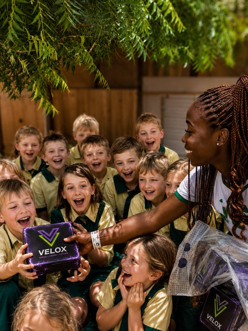 A group of excited school children in green and gold uniforms gather around a smiling woman with braided hair who is handing them a purple box labeled 'VELOX'.