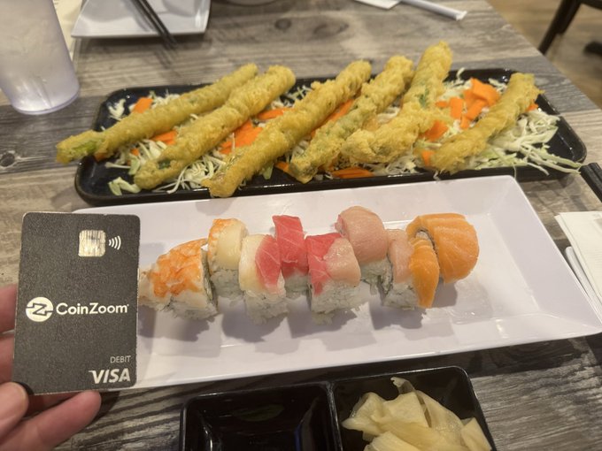A person holds a black CoinZoom VISA debit card in front of a meal featuring a plate of assorted sushi rolls and a tray of tempura.
