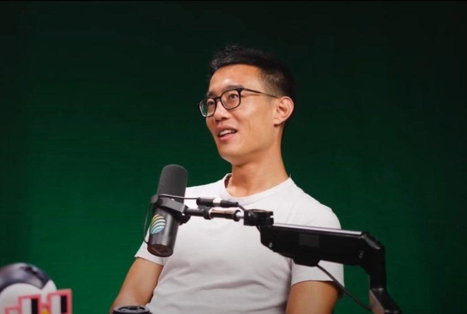 An Asian man wearing glasses and a white t-shirt speaks into a microphone against a green screen background.