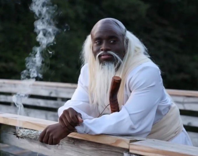 A bald Black man with a long white beard, dressed in white robes, leans on a wooden railing while holding a smoking pipe and bundle of herbs.
