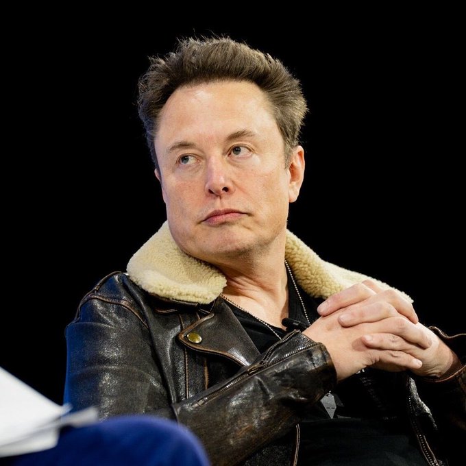 Elon Musk sits with hands clasped, gazing off thoughtfully while wearing a shearling-lined leather jacket. His contemplative expression visually echoes the post's quote about his priorities being driven by problem-solving rather than financial gain.