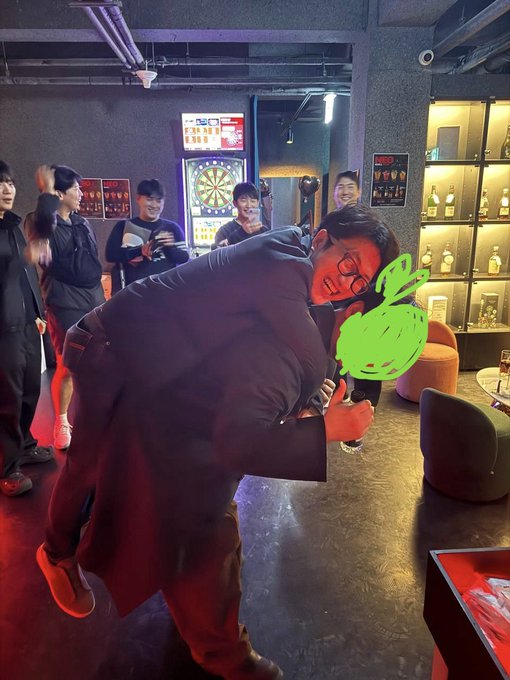 A man in glasses is being lifted up playfully by another person in a casual indoor setting that appears to be a bar or game room with a dartboard visible in the background, while other people smile and watch. One face is obscured with a green digital drawing.