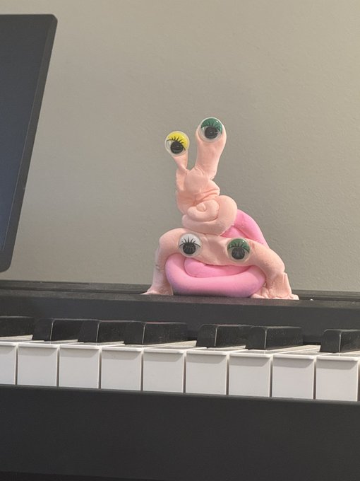 A pink sculpted creature with multiple googly eyes on stalks sits behind a laptop keyboard, peeking over the screen.