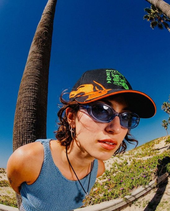 Selfie of a woman wearing sunglasses and a black and orange 'Harry Potter Slytherin' trucker hat, standing outdoors next to a palm tree under a blue sky.