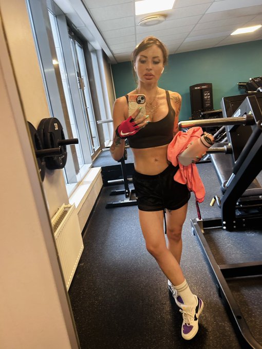 A mirror selfie of a woman in a gym wearing a black sports bra, shorts, and sneakers, holding a phone with a red case while wearing pink and orange gloves.
