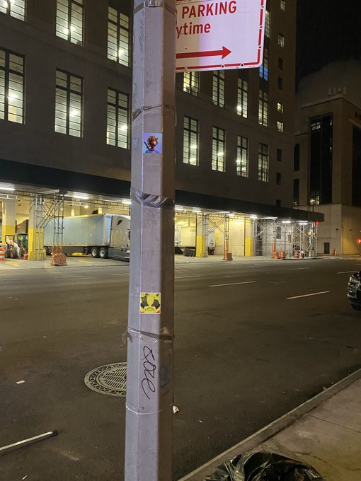 A nighttime urban street scene featuring a 'No Parking Anytime' sign on a metal pole covered in stickers, with a large truck parked in front of an illuminated brick building.