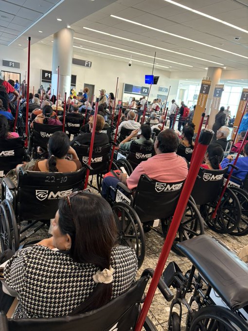 A crowded hospital waiting area filled with people sitting in wheelchairs that have 'MGE' and 'F-05' labels, with gate signs like 'F1' visible overhead.