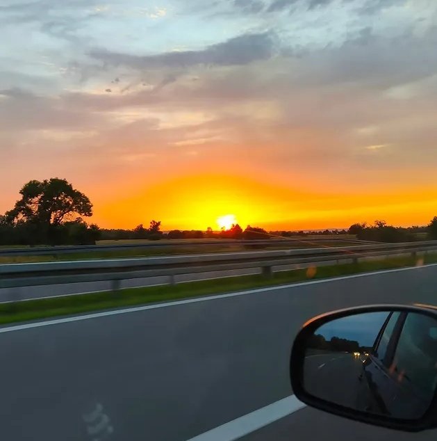 A car's rearview mirror reflects a sunset over a lush green field, with the sun setting behind trees and casting warm hues across the sky.